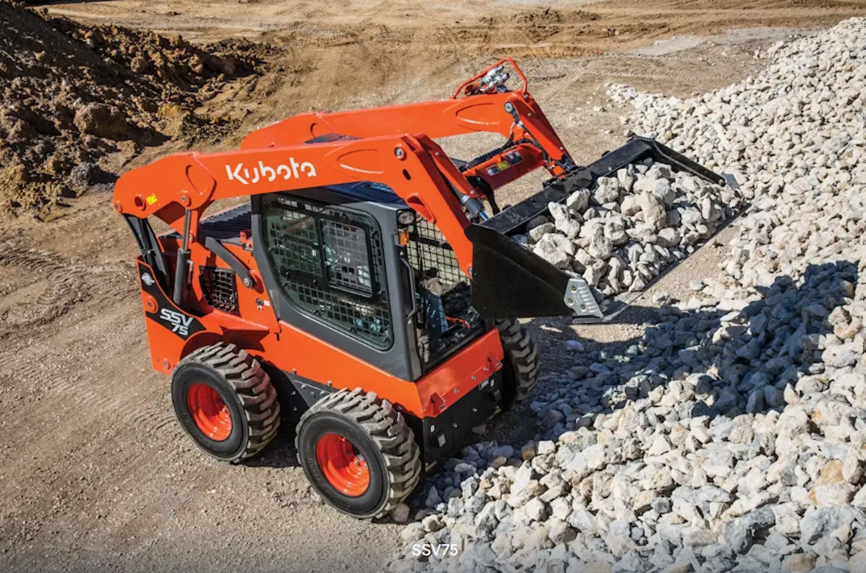 Orange Kubota SSV75 skid-steer loader scooping a pile of rocks with a bucket at a construction site