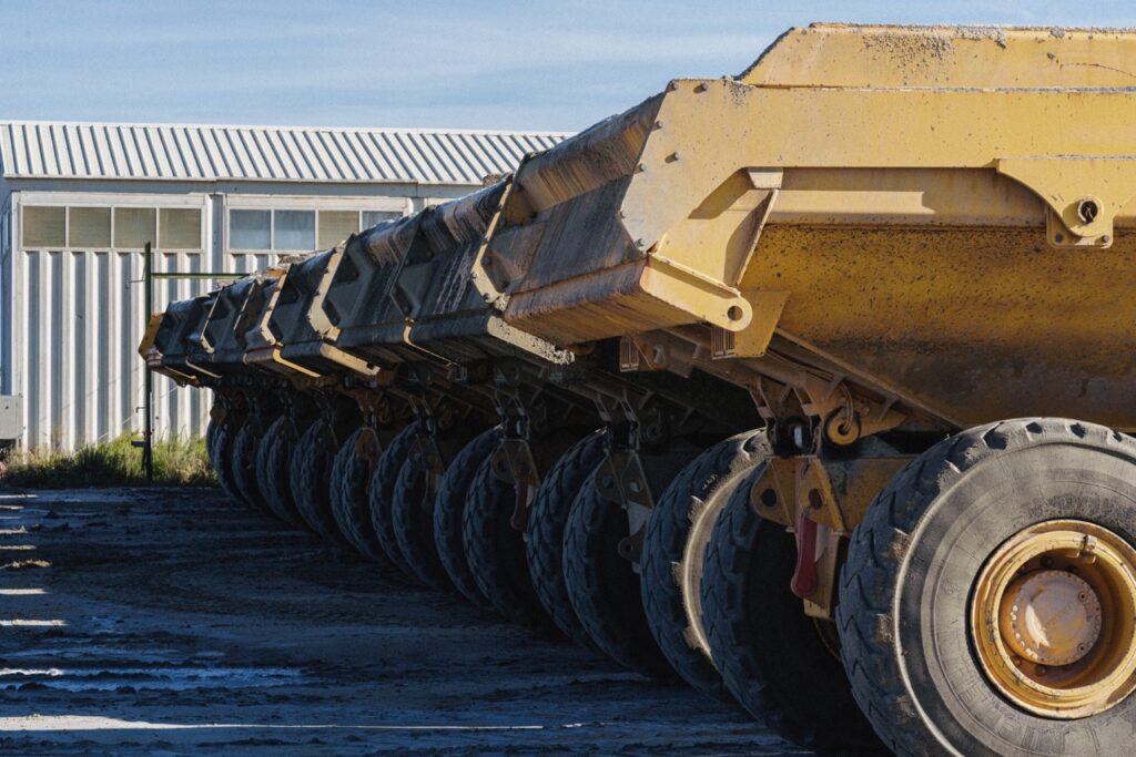 Row of large yellow dump trailer wheels and chassis parked beside a metal-sided building in a yard.
