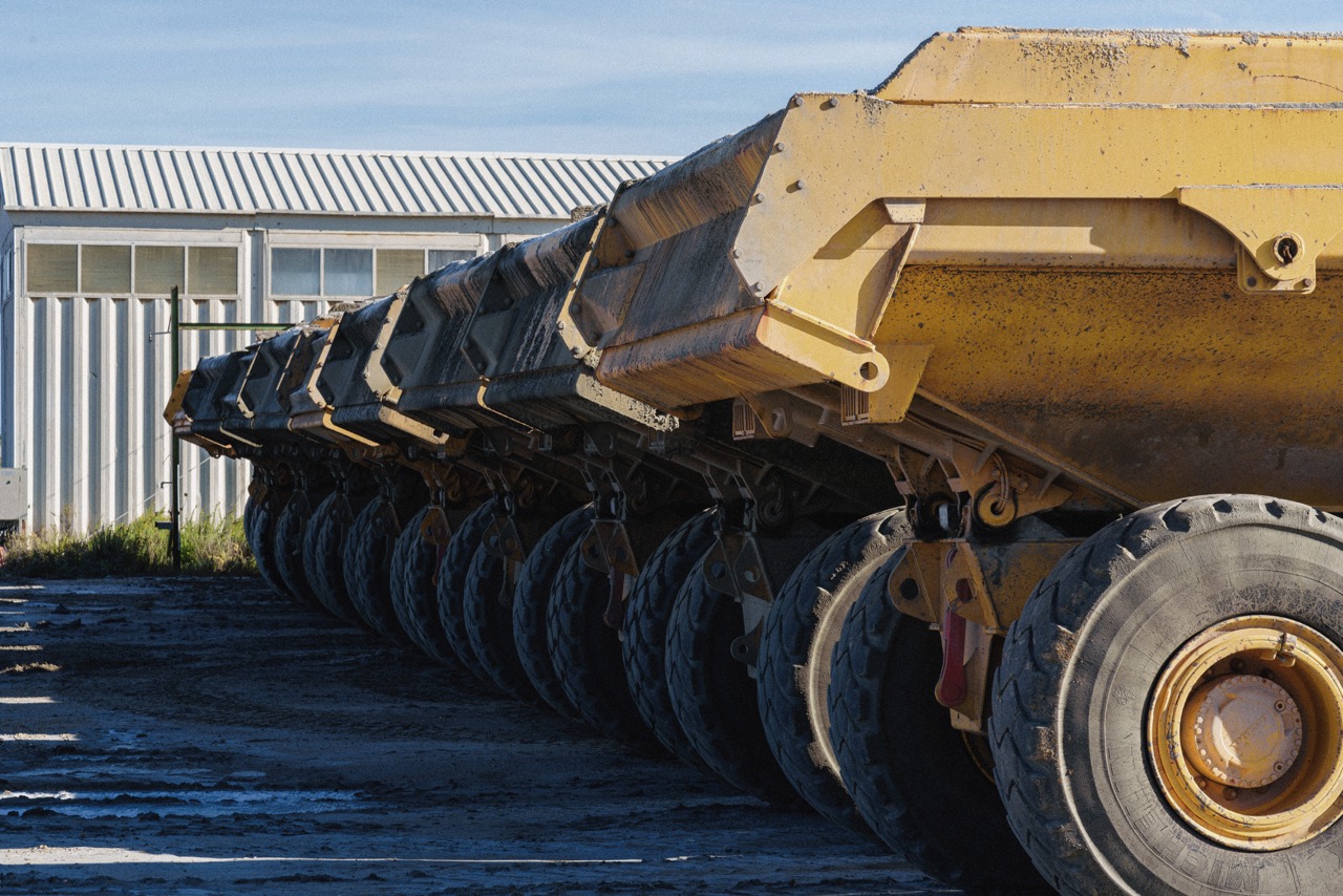 Row of large yellow dump trailer wheels and chassis parked beside a metal-sided building in a yard.