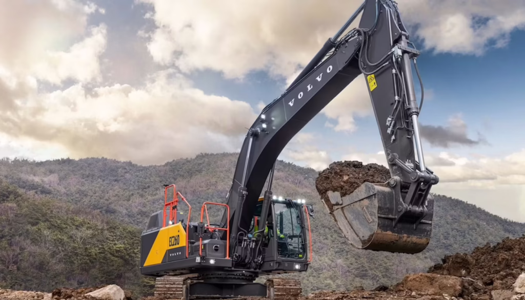 Volvo hydraulic excavator scooping soil at a rocky construction site with forested hills in the background