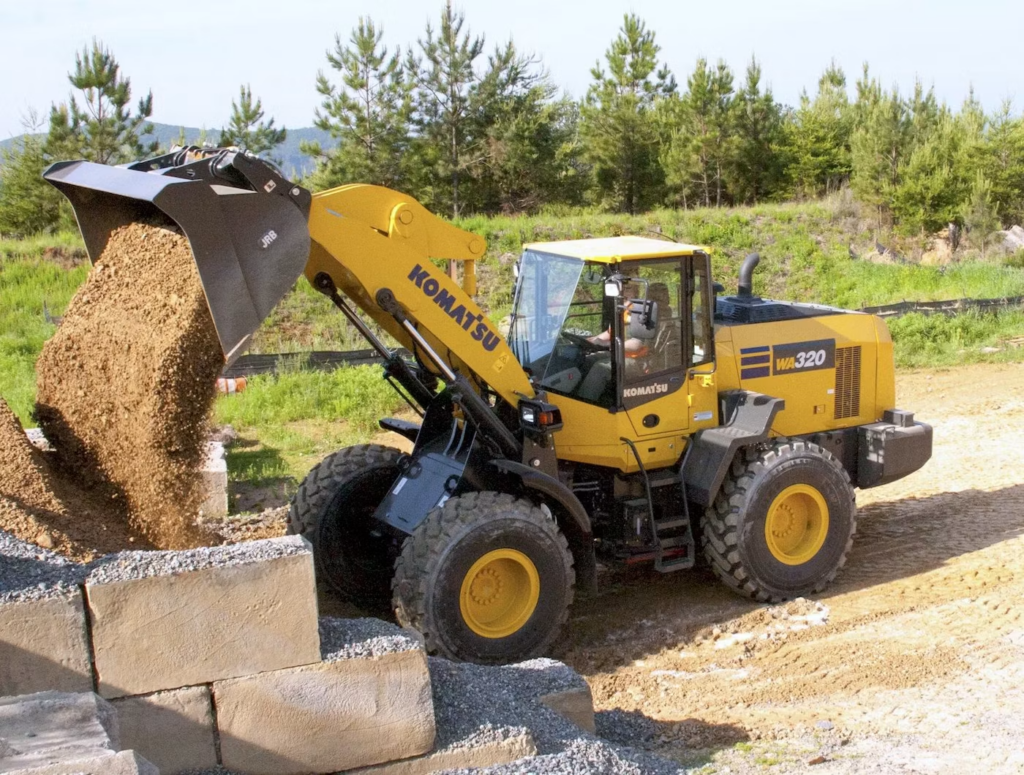 Yellow Komatsu WA320 wheel loader loading dirt from a pile at a construction site, with a concrete block wall in the foreground and trees in the background