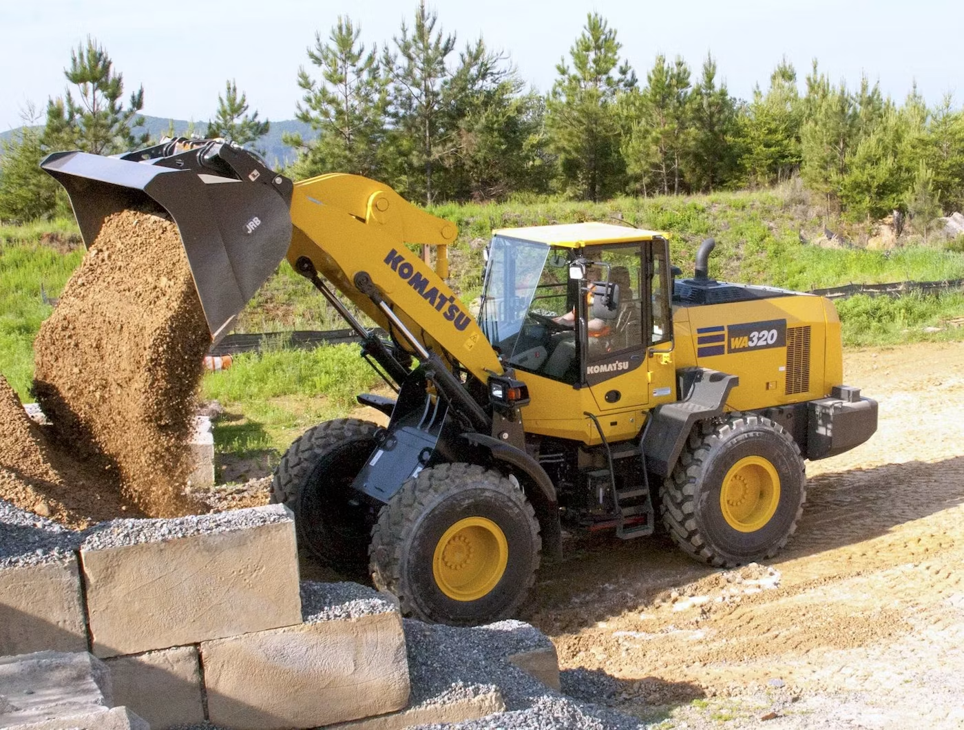 Yellow Komatsu WA320 wheel loader loading dirt from a pile at a construction site, with a concrete block wall in the foreground and trees in the background